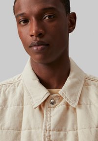 Giacca trapuntata beige con texture denim, dotata di colletto a punta e chiusura a pressione, abbinata a una camicia di colore chiaro sotto.