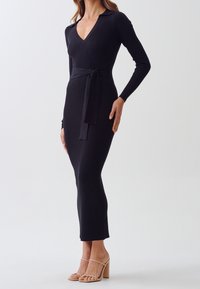 Svart ribbad klänning med V-ringning och långa ärmar, med bältesmidja och figursydd silhuett, som matchas med nude blockklacksandaler.