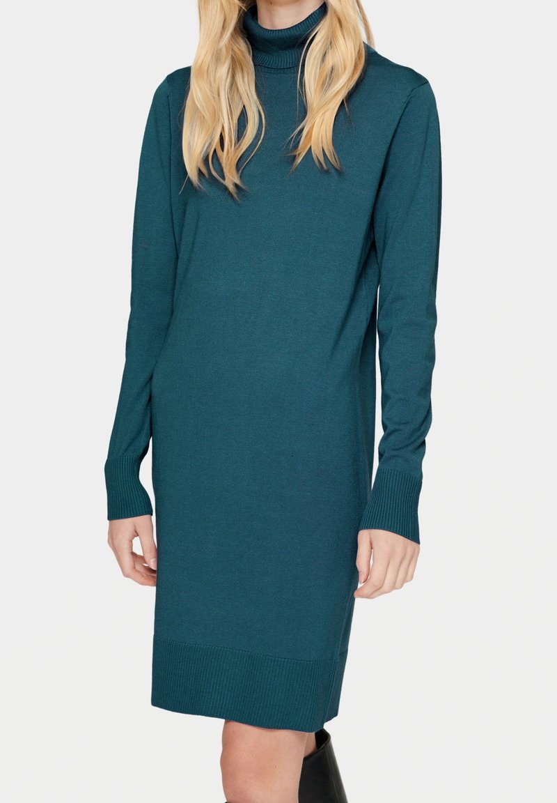 Femme portant une robe pull col roulé à manches longues avec poignets côtelés de couleur bleu sarcelle, associée à des bottes noires montant jusqu'aux genoux.