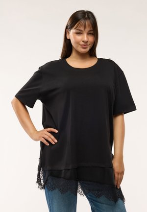 Giovane donna con lunghi capelli scuri che indossa una camicia nera larga con orlo in pizzo e jeans blu, mano sul fianco, su uno sfondo semplice e chiaro.