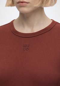 Gros plan d'une personne portant une chemise couleur rouille avec un col rond et un petit texte en relief "HUGO" sur la poitrine.