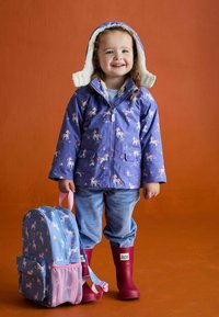 Lila wasserdichte Jacke mit Einhornmustern, gefütterte Kapuze aus Fleece, gestreifte blaue Hose, rote Gummistiefel und ein passender blauer Rucksack.