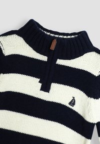 Strickpullover mit marineblauen und cremefarbenen horizontalen Streifen, mit einem hohen Kragen, einem halben Reißverschluss und einem kleinen gestickten Segelboot-Logo.