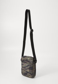 Piccola borsa a tracolla con motivo a camo astratto in nero, marrone e beige, e tracolla nera regolabile, appesa su uno sfondo chiaro.