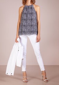 Haut halter à motifs noir et blanc avec des plis, associé à un jean skinny blanc. Une veste blanche est tenue d'une main. Sandales à talons argentés.