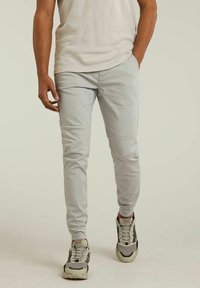 Pantalon fuselé gris clair en tissu lisse, avec une coupe slim, des détails en panneaux, porté avec des baskets grises et noires.