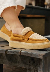 Yellow suede slip-on shoes with a fluffy cream fleece upper, featuring a strap across the foot and a textured cream sole.