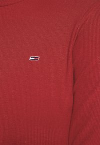 Roter rippenstrukturierter Pullover aus Stoff mit einem kleinen rechteckigen gestickten Logo in Weiß, Marineblau und Rot auf der Brust.