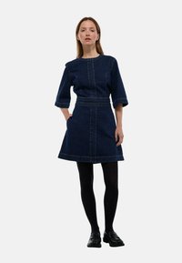 Femme portant une robe en denim bleu foncé à manches trois-quarts, des collants noirs et des chaussures noires, se tenant avec une main dans la poche sur un fond blanc.