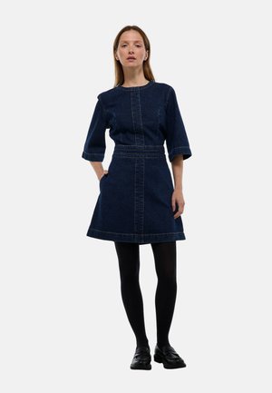 Femme portant une robe en denim bleu foncé à manches trois-quarts, des collants noirs et des chaussures noires, se tenant avec une main dans la poche sur un fond blanc.