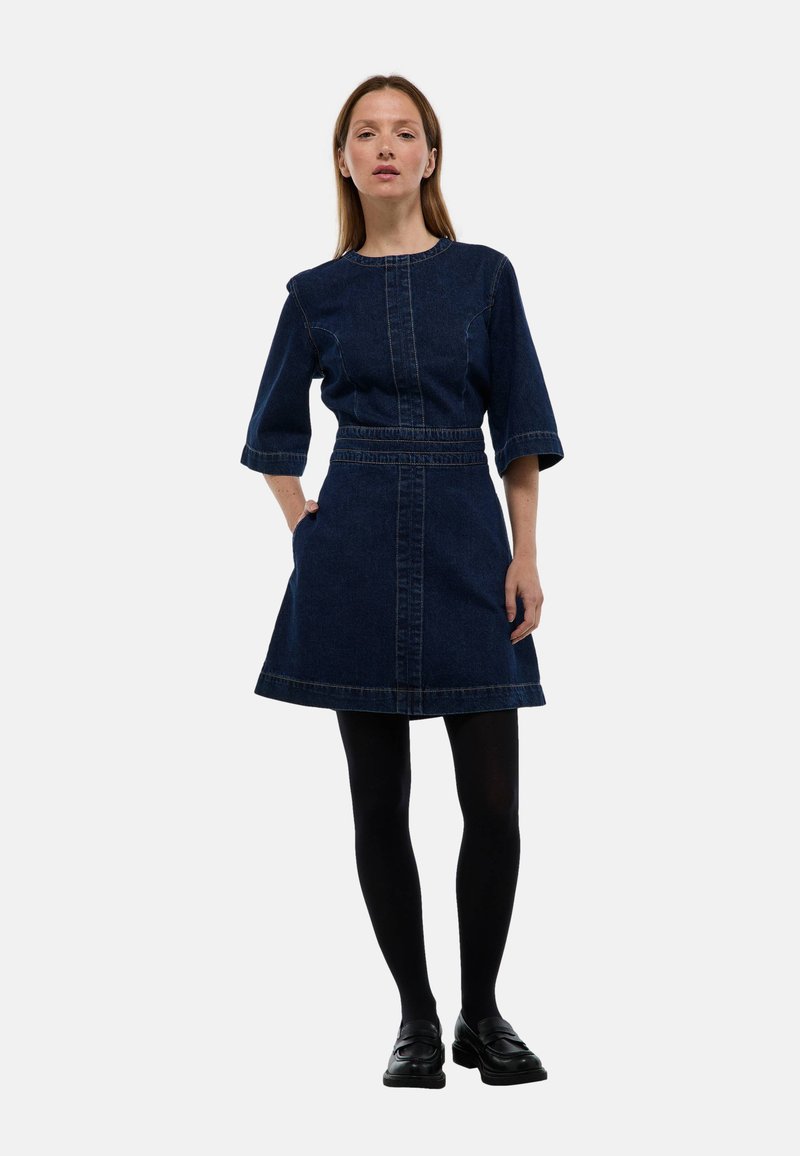 Femme portant une robe en denim bleu foncé à manches trois-quarts, des collants noirs et des chaussures noires, se tenant avec une main dans la poche sur un fond blanc.