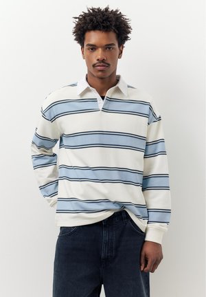 Jonge man met krullend haar, gekleed in een wit en blauw gestreept shirt met lange mouwen en donkere spijkerbroek, staand tegen een effen witte achtergrond.