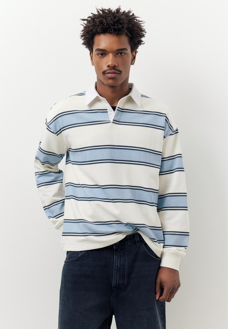 Jonge man met krullend haar, gekleed in een wit en blauw gestreept shirt met lange mouwen en donkere spijkerbroek, staand tegen een effen witte achtergrond.