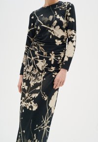 Mujer vistiendo un vestido negro de manga larga con patrones florales abstractos en color beige, ajustado a la cintura y que fluye en una falda larga.