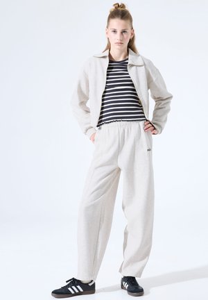 Lichtgrijs jack met ritssluiting, zwart-wit gestreept shirt, lichtgrijze broek met elastische tailleband, en zwarte sneakers met witte strepen.