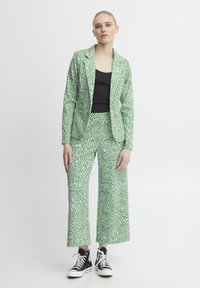 Green patterned blazer and matching wide-leg trousers with diamond shapes. Worn over a black top and paired with black sneakers.