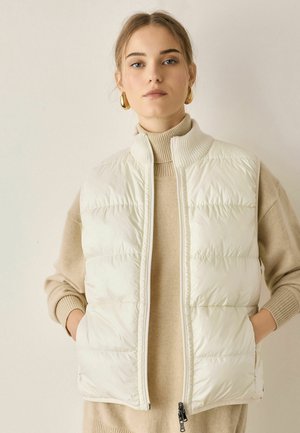 Kvinde iført en hvid quiltet ærmeløs vest over en beige rullekravesweater, med guldringearringe, stående mod en ensfarvet baggrund.