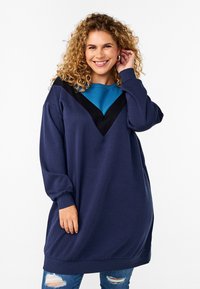 Abito felpa oversize color navy con un design a zigzag teal e nero. Tessuto morbido, polsini a costine e orlo arrotondato. Leggera usura sui jeans.