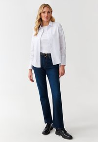 Hvid skjorte med knapper over en hvid top, parret med mørkeblå flagrende jeans og sorte sko. Har et bælte med en rund spænde.