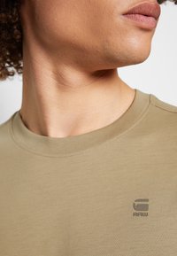 Gros plan d'une personne portant un t-shirt beige avec un petit logo "G RAW" sur la poitrine gauche, regardant sur le côté.
