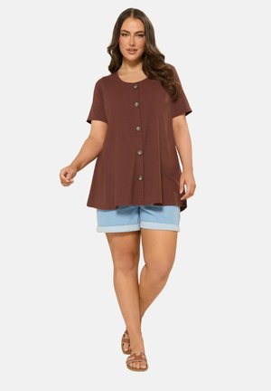 Kvinde iført en brun, knappet kortærmet top, lyseblå op rullede denimshorts og brune flade sandaler, står foran en ensfarvet baggrund.