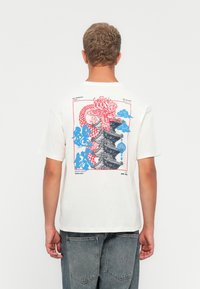 Cream-colored cotton t-shirt featuring a large red and blue dragon graphic and pagoda design on the back. Relaxed fit with short sleeves.