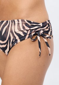 Κοντινό πλάνο ενός ατόμου που φοράει μπικίνι με μοτίβο ζέβρας και κόμπο στο πλάι στο ισχίο, με απλό φόντο.