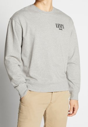 Mężczyzna ubrany w jasnoszarą bluzę Levi’s z logo na piersi i beżowe spodnie, stojący z jedną ręką w kieszeni na jednolitym tle.