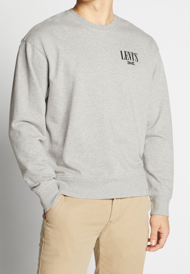 Homme portant un sweat-shirt Levi’s gris clair avec un logo sur la poitrine et un pantalon beige, debout avec une main dans la poche sur un fond uni.