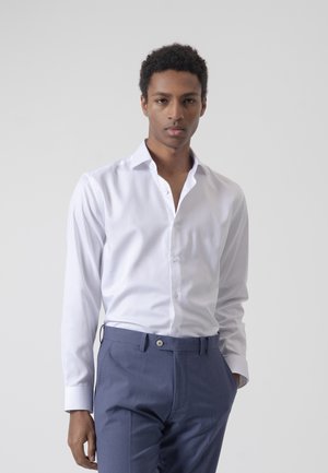 Joven con una camisa blanca ajustada y pantalones azules, de pie frente a un fondo liso de color gris claro.