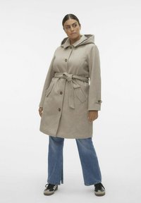 Abrigo de lana color beige claro con capucha, cierre de botones, cinta en la cintura y dos bolsillos delanteros; combinado con pantalones de mezclilla azul de corte acampanado y zapatillas negras.