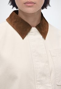 Γυναίκα που φοράει μπεζ πουκάμισο με καφέ σουέτ γιακά και μπροστινή τσέπη, εμφανιζόμενη από το λαιμό έως το άνω μέρος του στήθους.