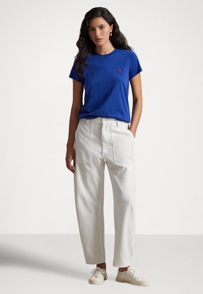 Kvinne med langt mørkt hår iført en kongeblå kortermet skjorte, hvite høytlivsbukser og hvite joggesko, stående med den ene hånden i lommen.