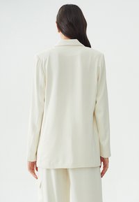 Crème-kleurige blazer met een glad, op maat gemaakt ontwerp, reverskraag en lange mouwen. Heeft een rechte snit en subtiele details aan de achterkant.