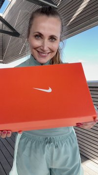 Oranžna Nike škatla za čevlje z belim logotipom, ki jo drži oseba v svetlo modri obleki. Delno vidna je lesena terasa v ozadju.