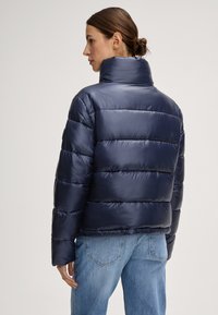 Giacca imbottita blu navy con trapuntatura verticale, colletto alto e una texture liscia e lucida; abbinata a jeans in denim azzurro.