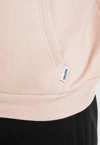 Sweatshirt rose clair avec ourlets et poignets côtelés, présentant une poche avant et une petite étiquette de marque sur le côté. Texture de tissu douce.
