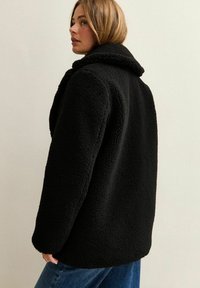 Mujer con cabello rubio corto vestida con una chaqueta de forro polar texturizada negra y jeans azules, de pie contra una pared clara y lisa.