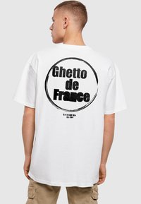 Balta medvilninė marškinėliai su dideliu apvaliu piešiniu ant nugaros, su juodu tekstu: "Ghetto de France" ir "Ça vient de la rue."