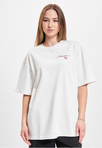 White oversized T-shirt made of cotton. Features short sleeves and a round neckline. Red embroidered text on the left chest area.