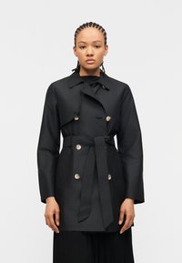 Γυναίκα με πλεγμένα μαλλιά, φορώντας μαύρο διπλής σειράς παλτό με ζώνη και μεγάλα κουμπιά, στέκεται μπροστά σε λευκό φόντο.