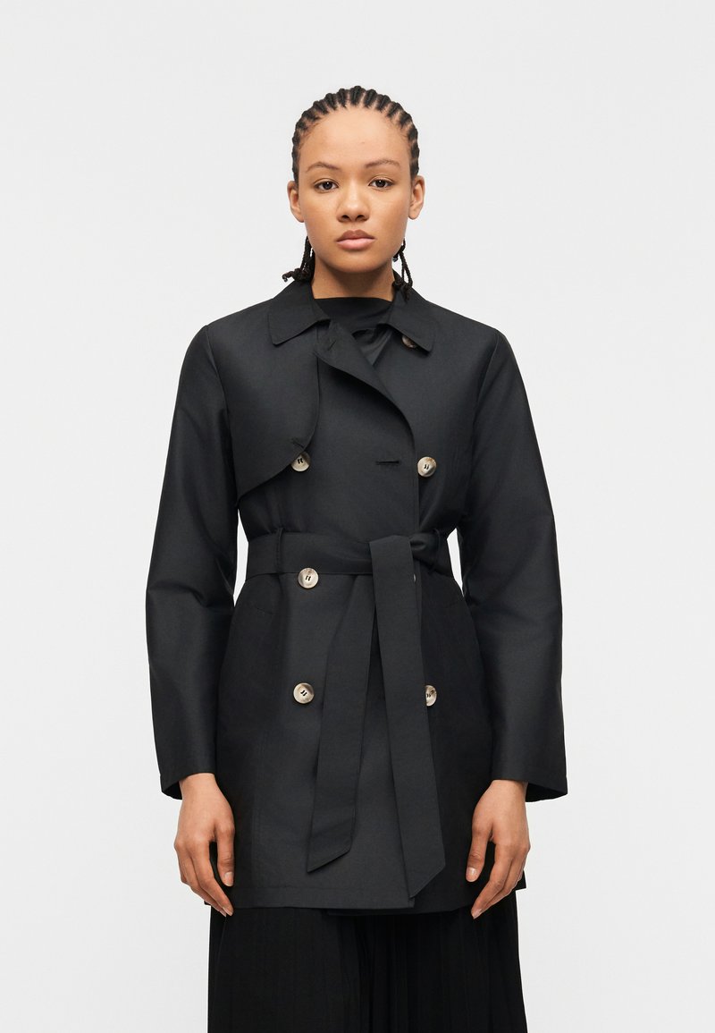 Γυναίκα με πλεγμένα μαλλιά, φορώντας μαύρο διπλής σειράς παλτό με ζώνη και μεγάλα κουμπιά, στέκεται μπροστά σε λευκό φόντο.