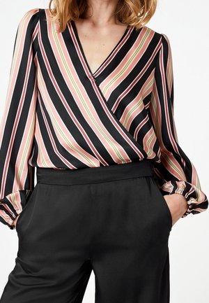 Femme portant un chemisier à manches longues et col en V avec des rayures diagonales roses, crème et noires, rentrée dans un pantalon noir taille haute, les mains dans les poches.