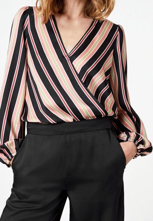 Femme portant un chemisier à manches longues et col en V avec des rayures diagonales roses, crème et noires, rentrée dans un pantalon noir taille haute, les mains dans les poches.