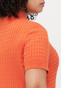 Oranje trui met korte mouwen, voorzien van een textuur met kabelgebreide patronen. Heeft een ronde halslijn en ribgebreide manchetten aan de mouwen.