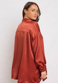Young woman with long brown hair wearing a loose shiny rust-colored blouse, looking over her shoulder against a white textured wall.