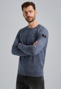 Blauer Strickpullover mit Rundhalsausschnitt, Raglanärmeln und strukturiertem Finish. Verfügt über ein kleines schwarzes Logopatch am linken Ärmel. Slim Fit.