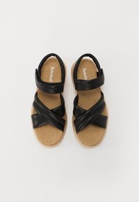 Sandales en cuir noir avec des brides entrecroisées, une semelle intérieure rembourrée beige, et une semelle extérieure texturée beige. Le logo est visible sur la semelle intérieure.