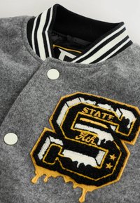 Šedá vlnená bomber bunda s čierno-bielym pruhovaným golierom, obsahuje veľké čierno-žlté vyšívané písmeno "S" a detail s kvapkami.