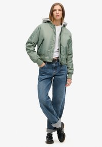 Grøn bomberjakke med grå hætte, lynlåslommer, ribbede manchetter, hvid t-shirt og opkrøllede blå jeans med sorte sko.